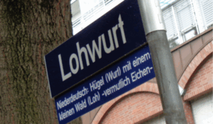 Straßenschild mit der Aufschrift "Lohwurt", darunter die Erklärung: "Niederdeutsch: Hügel (Wurt)mit einem kleinen Hügel (Loh) -vermutlich Eichen-"