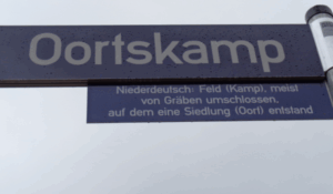 Straßenschild mit der Aufschrift "Oortskamp", darunter die Erklärung: "Niederdeutsch: Feld (Kamp), meist von Gräben umschlossen, auf dem eine Siedlung (Oort) entstand"